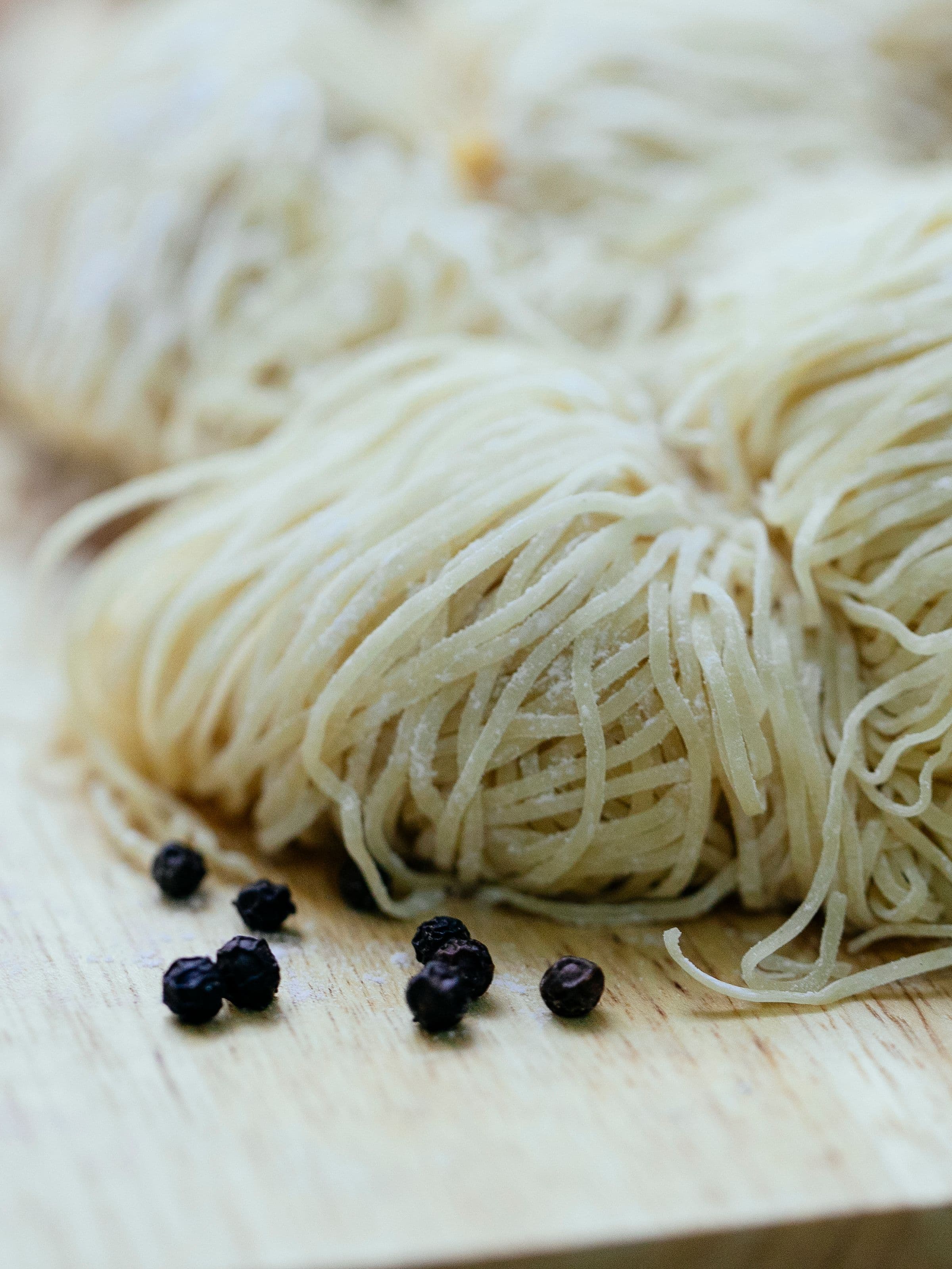 A nest of very fine, flour-dusted raw angel-hair strands on a warm wooden board, with black peppercorns for scale