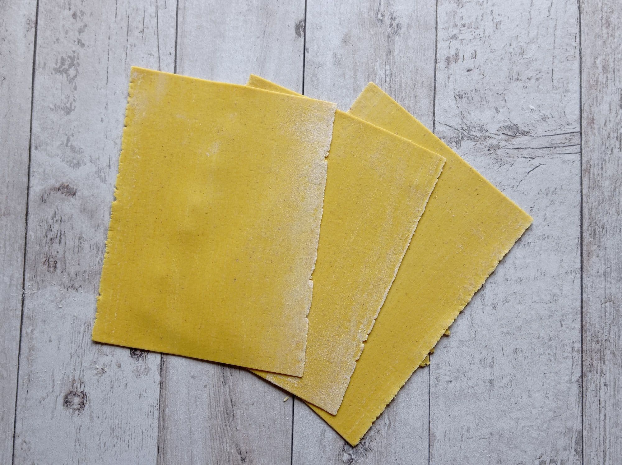 Wide flat sheets of raw yellow lasagna pasta fanned out on a rustic wooden board