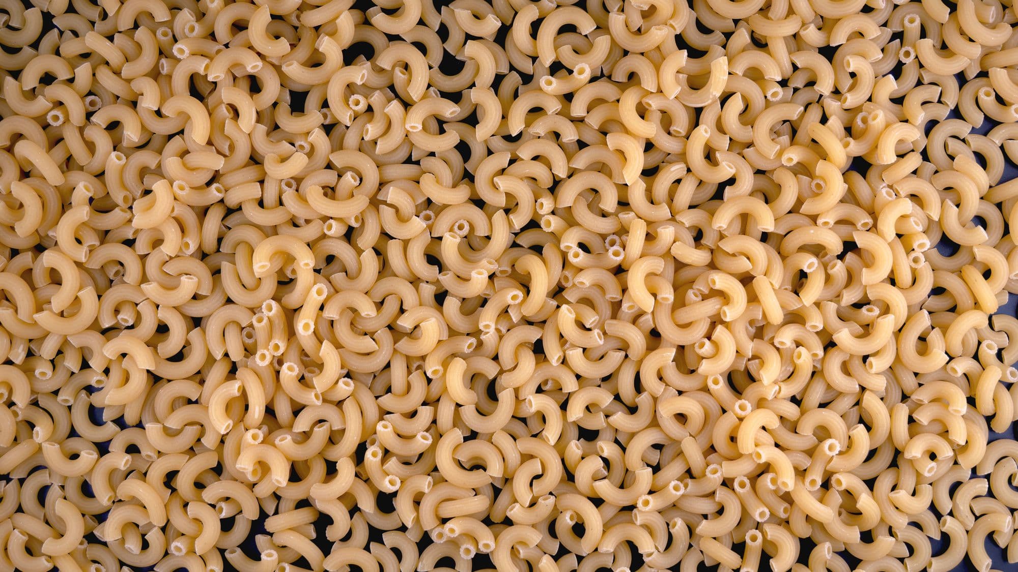 A fill-the-frame macro of raw elbow macaroni, each a ridged golden C-curve with hollow ends